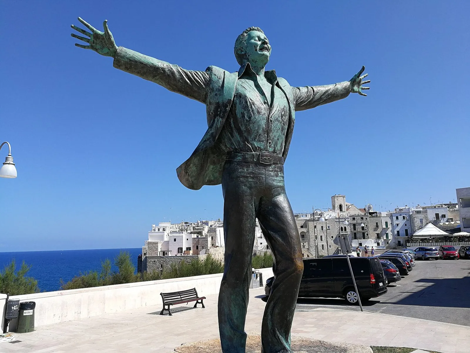 La statua in bronzo di Domenico Modugno a braccia aperte sul lungomare di Polignano a Mare, con il borgo antico e il mare sullo sfondo