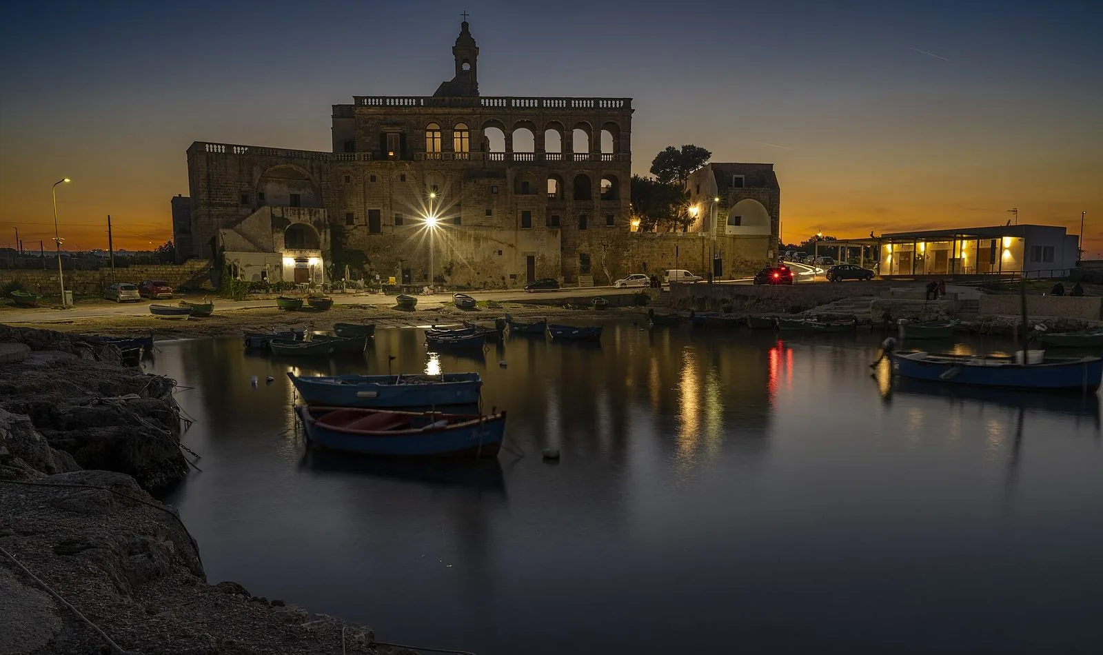 L'Abbazia di San Vito Martire a Polignano a Mare al crepuscolo, con le barche dei pescatori ormeggiate nel piccolo porto davanti alla facciata illuminata