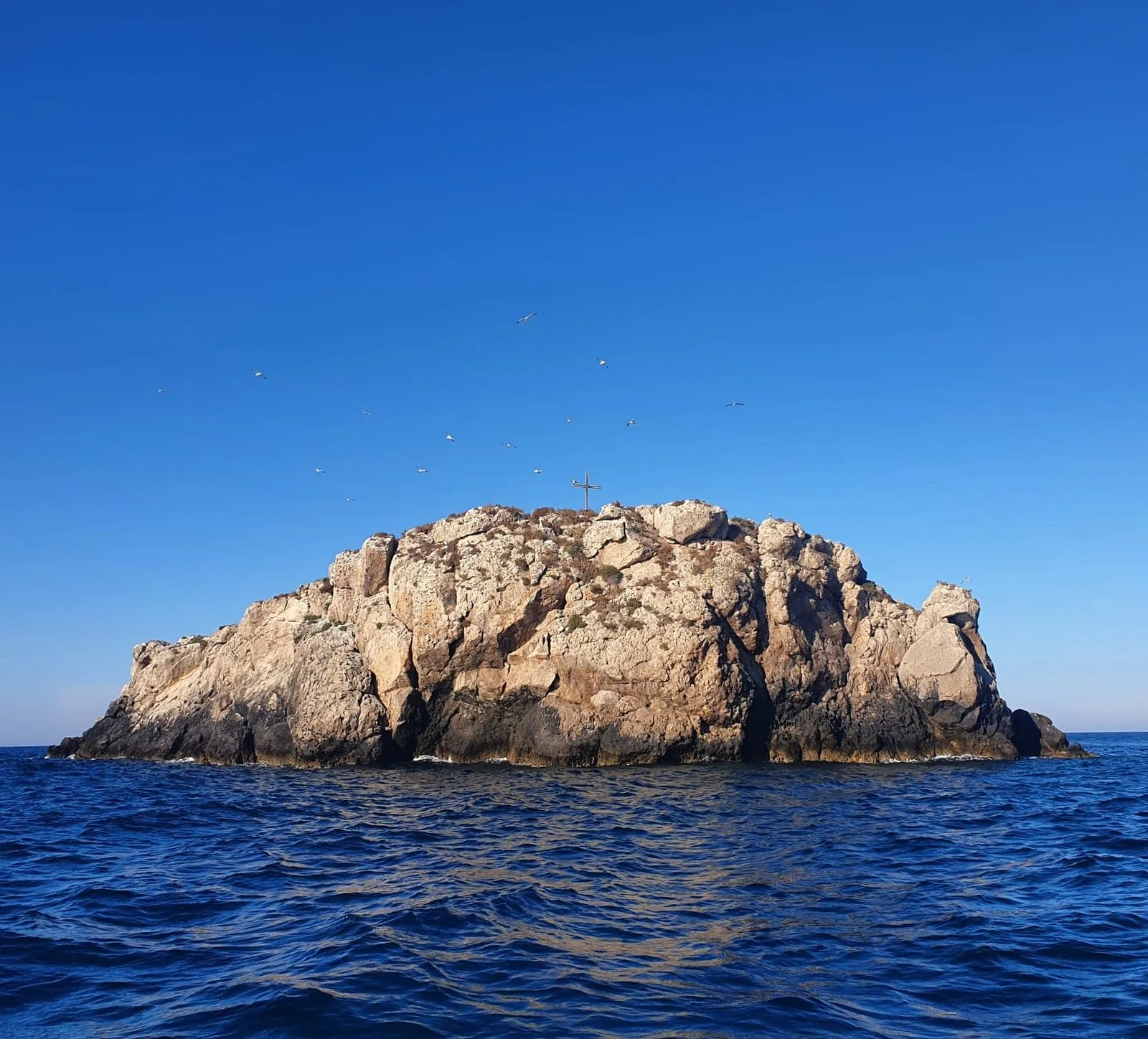 Lo Scoglio dell'Eremita, isolotto roccioso con una croce sulla sommità, visto dal mare di Polignano a Mare in una giornata di sole, con gabbiani in volo nel cielo azzurro