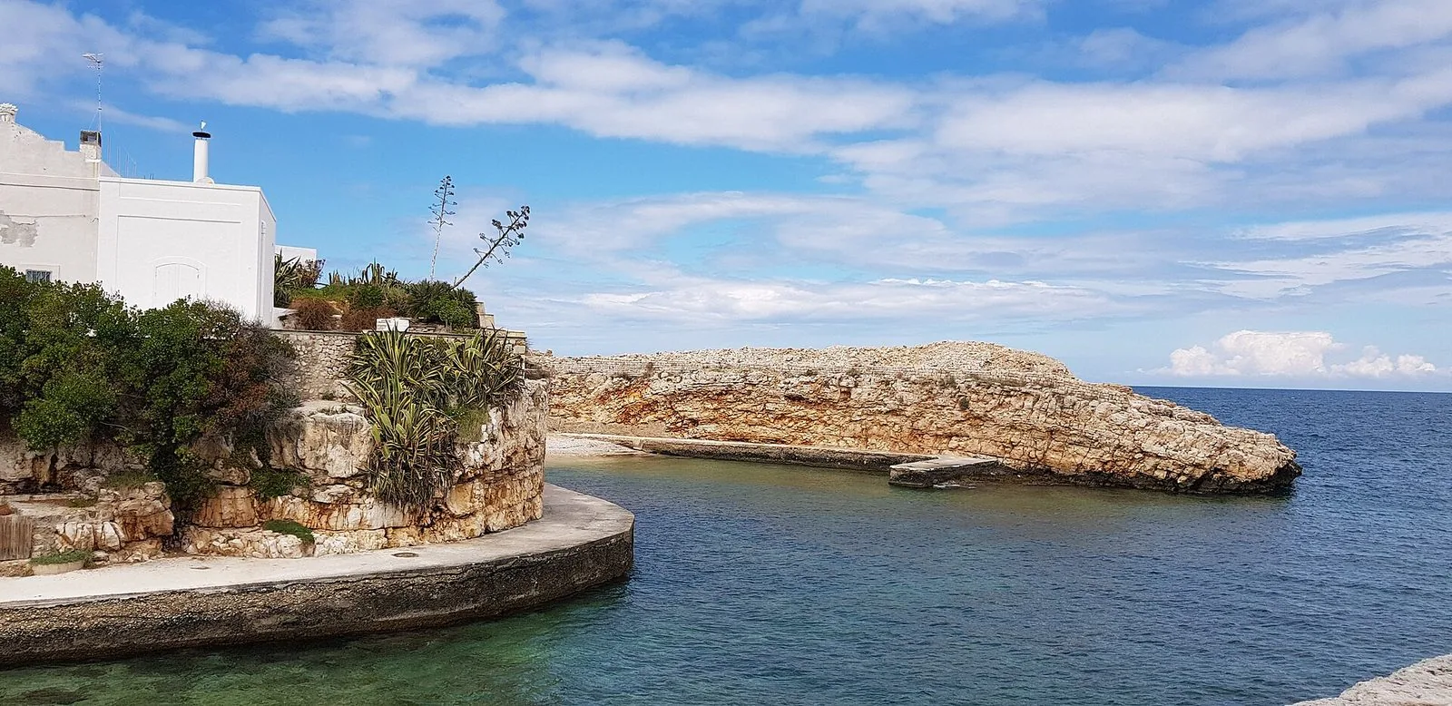 Cala Paura a Polignano a Mare: insenatura di acqua limpida con una passerella in cemento, uno sperone di roccia calcarea e una casa bianca tra agavi e vegetazione